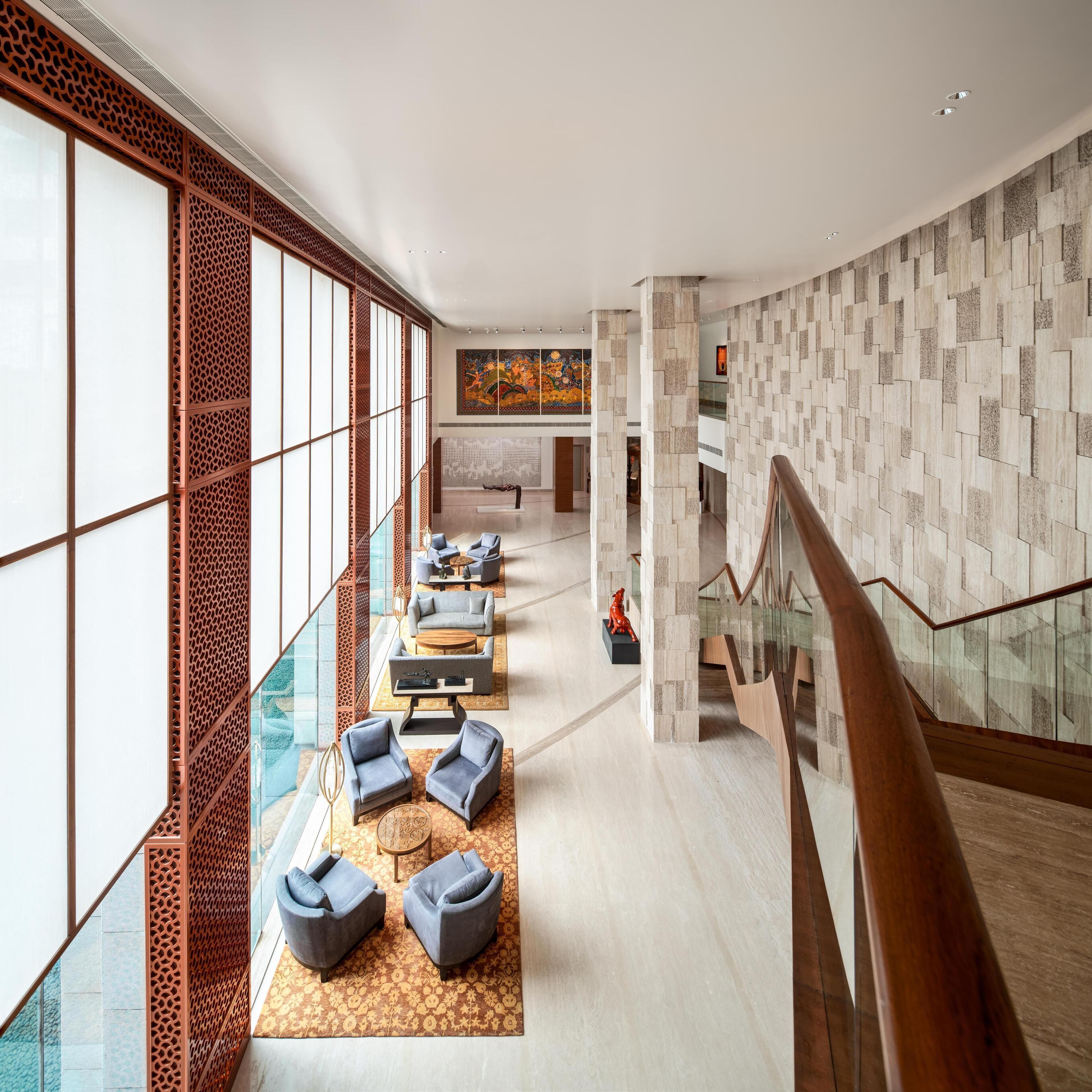 A spacious lobby with a modern design, featuring a seating area with sofas and a coffee table, a staircase with glass railings, and a decorative wall with a patterned design.