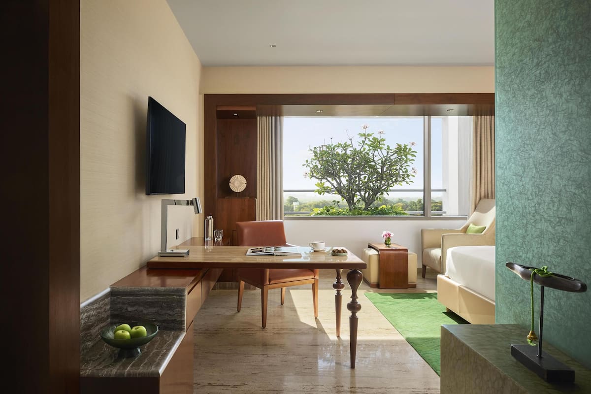 A modern hotel room with a dining area, a large window with a view of greenery, a flat-screen TV, and a bed with a green accent wall.