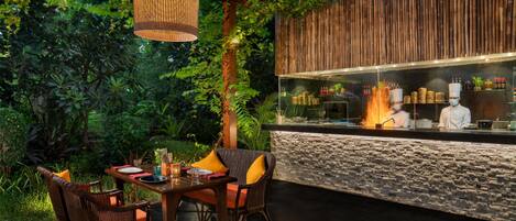 An outdoor dining area with wicker chairs and a table, a chef at the counter, and a stone wall backdrop.