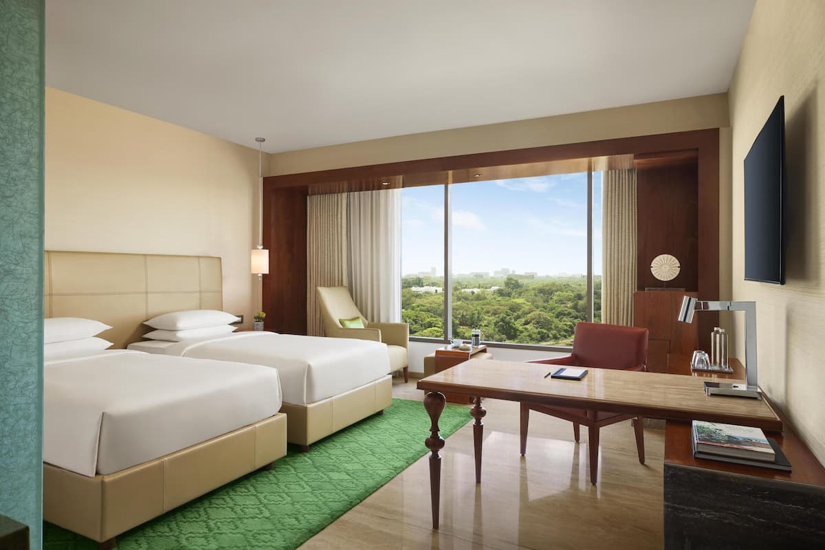 A hotel room with two beds, a desk, a chair, a TV, and a large window with a city view.