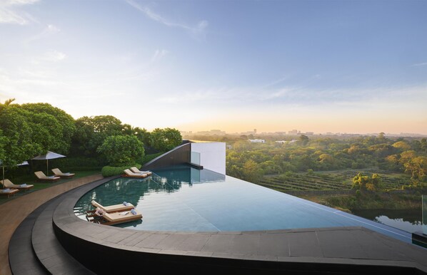 Ein moderner Infinity-Pool mit gewölbter Kante, umgeben von Liegestühlen und Sonnenschirmen, mit Blick auf eine üppige Landschaft und eine entfernte Stadtlandschaft.