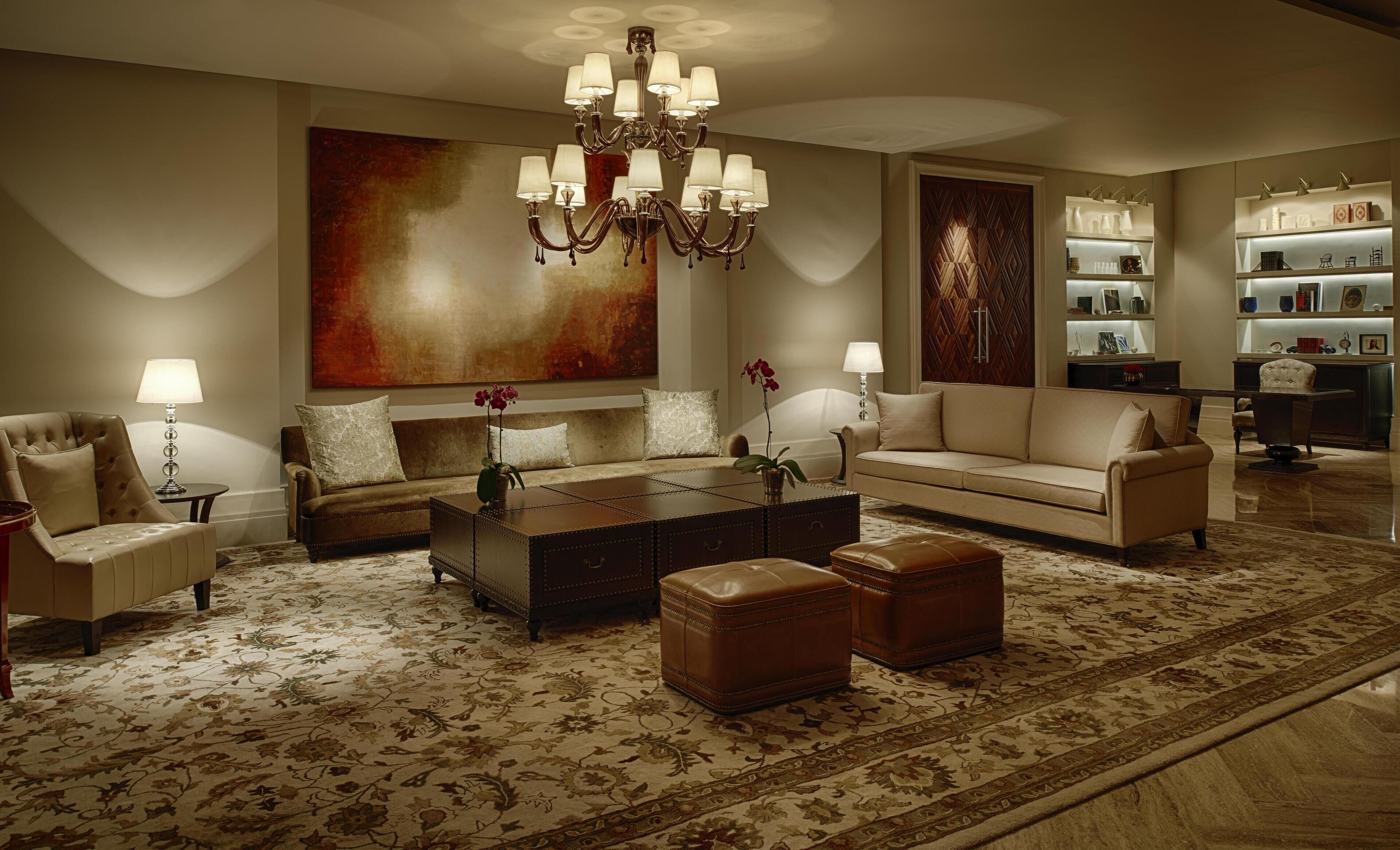 A spacious living room with a large sofa, a coffee table, and a chandelier. There is a bookshelf with decorative items and a large painting on the wall.