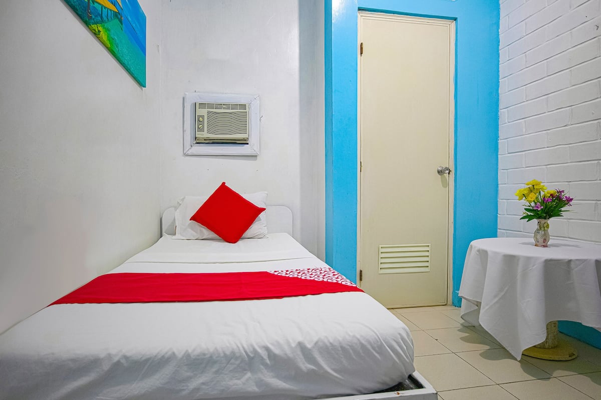 A bedroom with a bed, a red pillow, a white blanket, a small table with a vase of flowers, and a door with a blue frame.