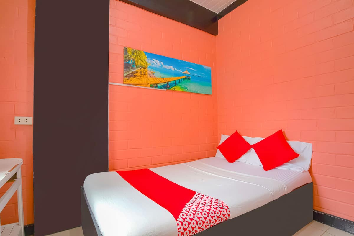 A single bed with a red and white patterned bedspread, two red pillows, and a painting of a tropical scene on the orange brick wall.