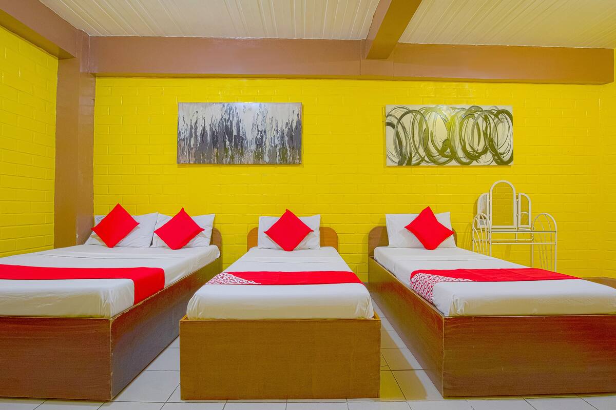 A hotel room with three beds, each with red and white bedding, and two paintings on the yellow brick wall.