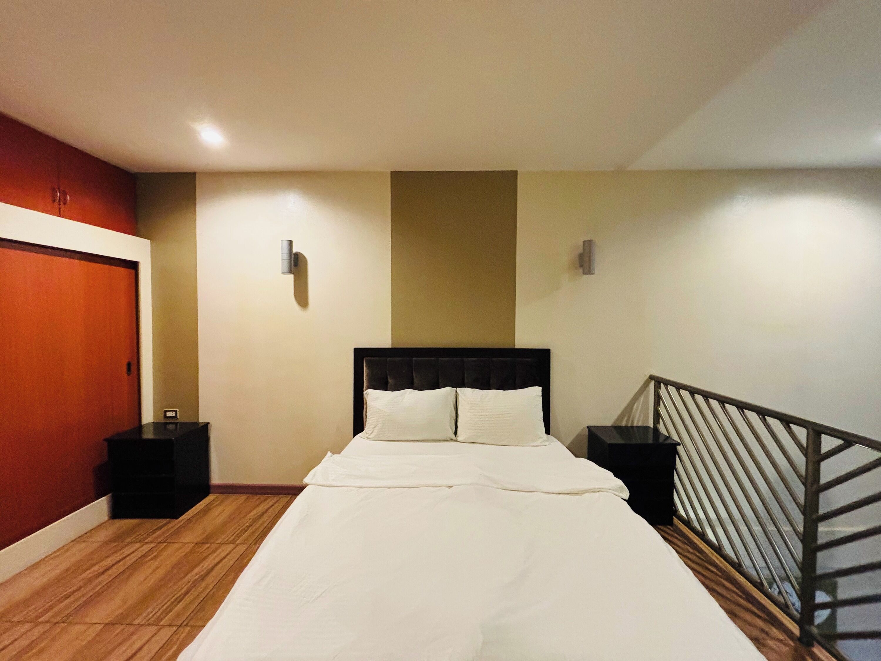 A hotel room with a bed, nightstands, a wooden floor, a red door, and a staircase.