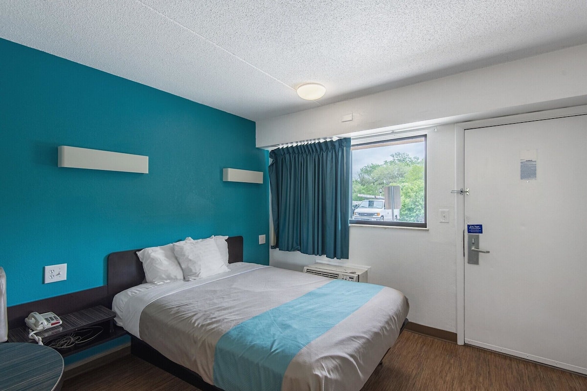 Une chambre d’hôtel avec un lit, un bureau, un téléphone, une fenêtre avec des rideaux et une porte avec une pancarte.