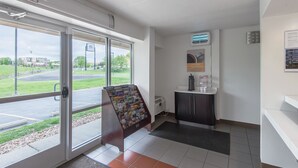 A hotel lobby with a reception desk, a coffee machine, a bulletin board with travel brochures, and a view of the parking lot and building outside.