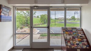 A hotel entrance with a glass door, a display of travel brochures, and a view of a green landscape with a parking lot.