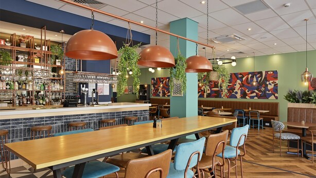A restaurant with a long wooden table, blue cushioned chairs, hanging pendant lights, and a bar area with shelves of bottles and glasses. - Travelodge London Woolwich (London)
