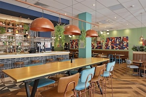 A restaurant with a long wooden table, blue cushioned chairs, hanging pendant lights, and a bar area with shelves of bottles and glasses. - Travelodge London Woolwich (London)