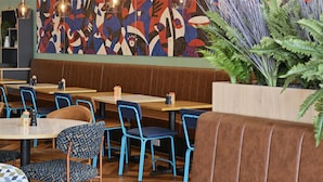 A restaurant interior with wooden tables, blue chairs, and a colorful abstract mural on the wall. - Travelodge London Woolwich (London)