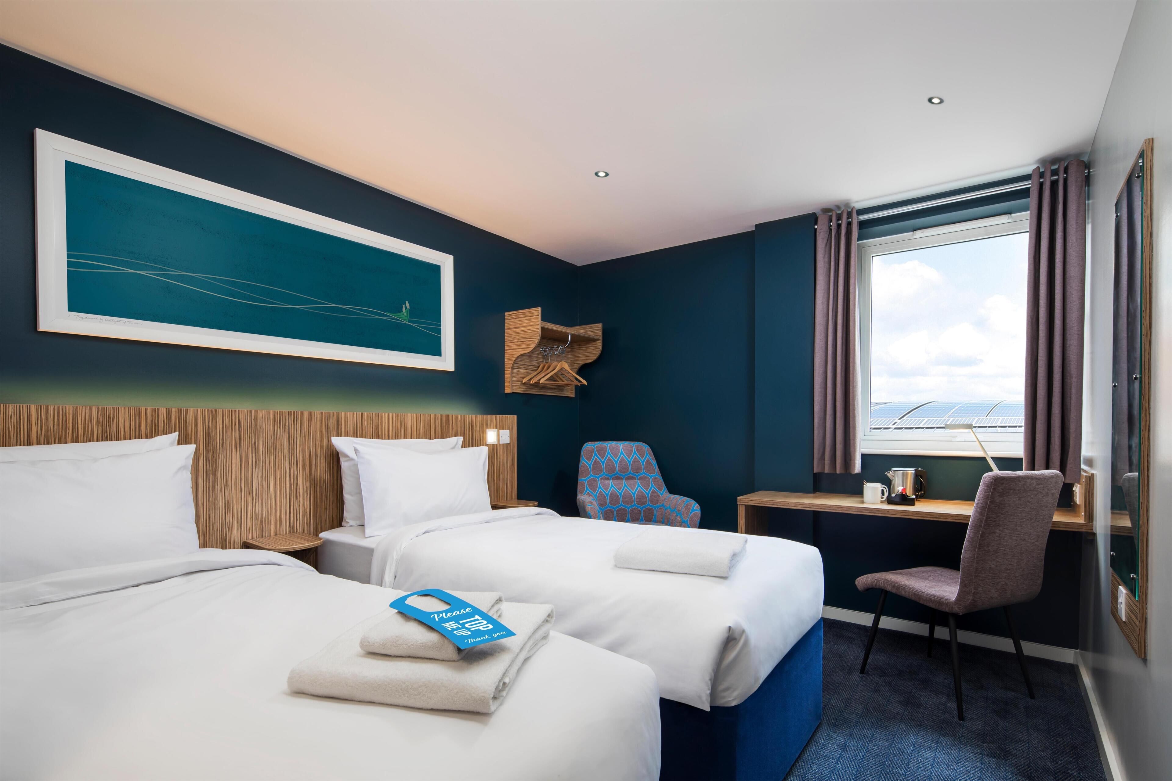 A hotel room with two beds, a desk, a chair, a large window, and a painting on the wall.