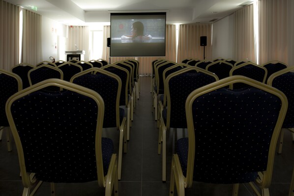 Un salón de conferencias con filas de sillas orientadas hacia una pantalla, un atril y cortinas.