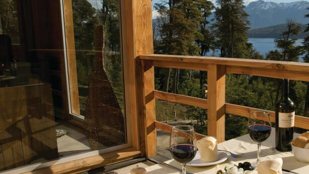 Una mesa con copas de vino, una botella y pan en un balcón con vistas a un lago y un bosque.