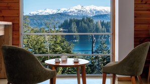 Habitación con un amplio ventanal que ofrece vistas a montañas nevadas, un lago y un bosque denso. En primer plano, una mesa redonda de madera con dos sillas.