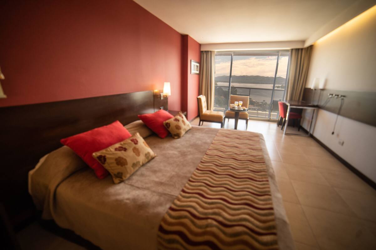 A hotel room with a large bed, a small table, a chair, and a view of the city through the window.
