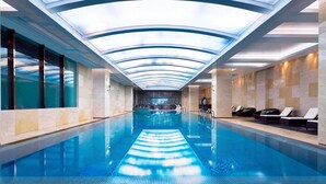 A modern indoor swimming pool with a glass ceiling, lounge chairs, and a view of the pool. - Sheraton Qingdao Jiaozhou Hotel (Qingdao)
