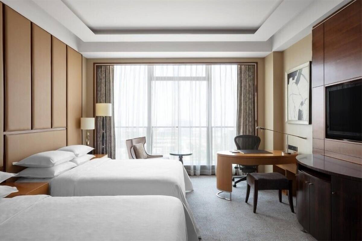 Ein Hotelzimmer mit zwei Betten, einem Schreibtisch, einem Sessel, einem Fernseher und einem großen Fenster.