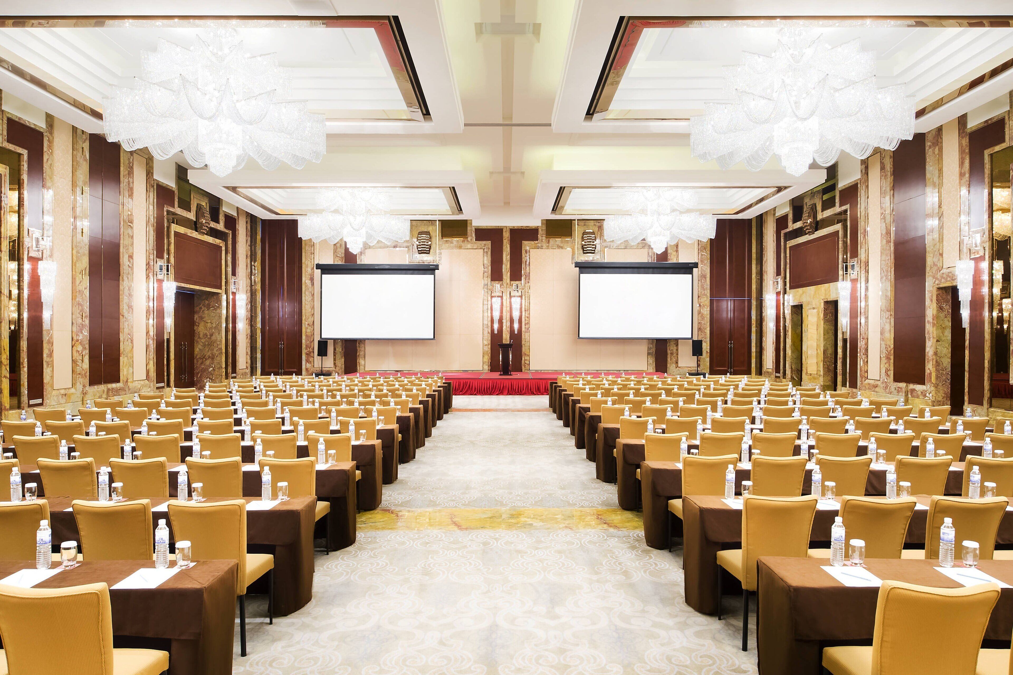A large conference room with rows of tables and chairs, a stage, and a large screen at the front.