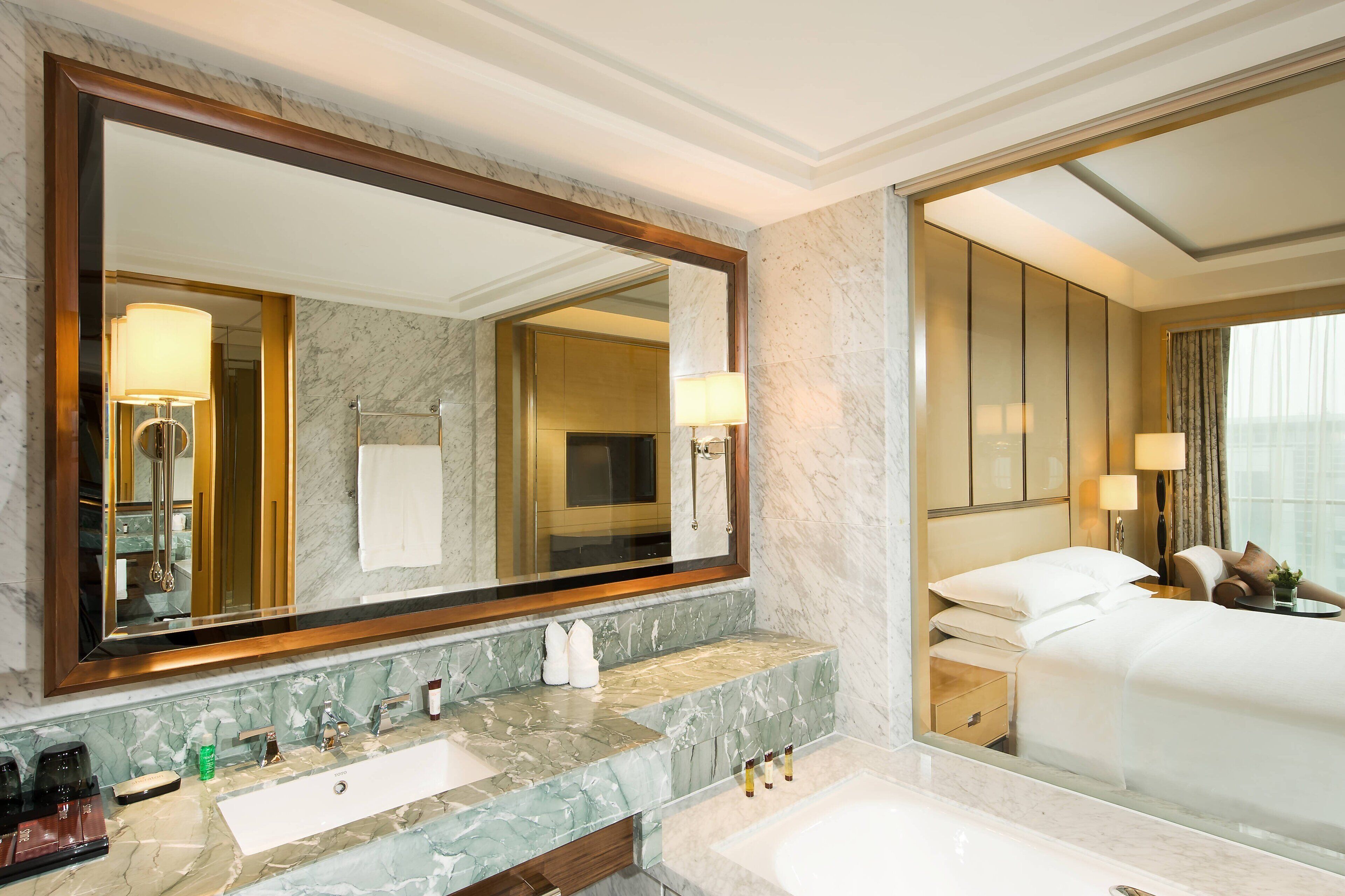 A hotel bathroom with a large mirror, marble countertop, sink, and a bed with a nightstand.