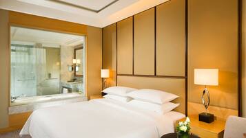 A hotel room with a large bed, a sitting area with a chair, a small table with a vase, and a large mirror.