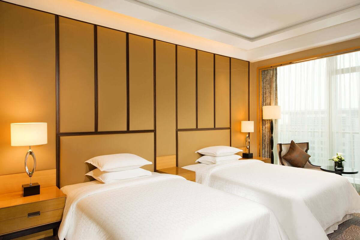 Ein Hotelzimmer mit zwei Betten, einem Nachttisch mit Lampe, einem Sessel und einem Fenster mit Vorhängen.