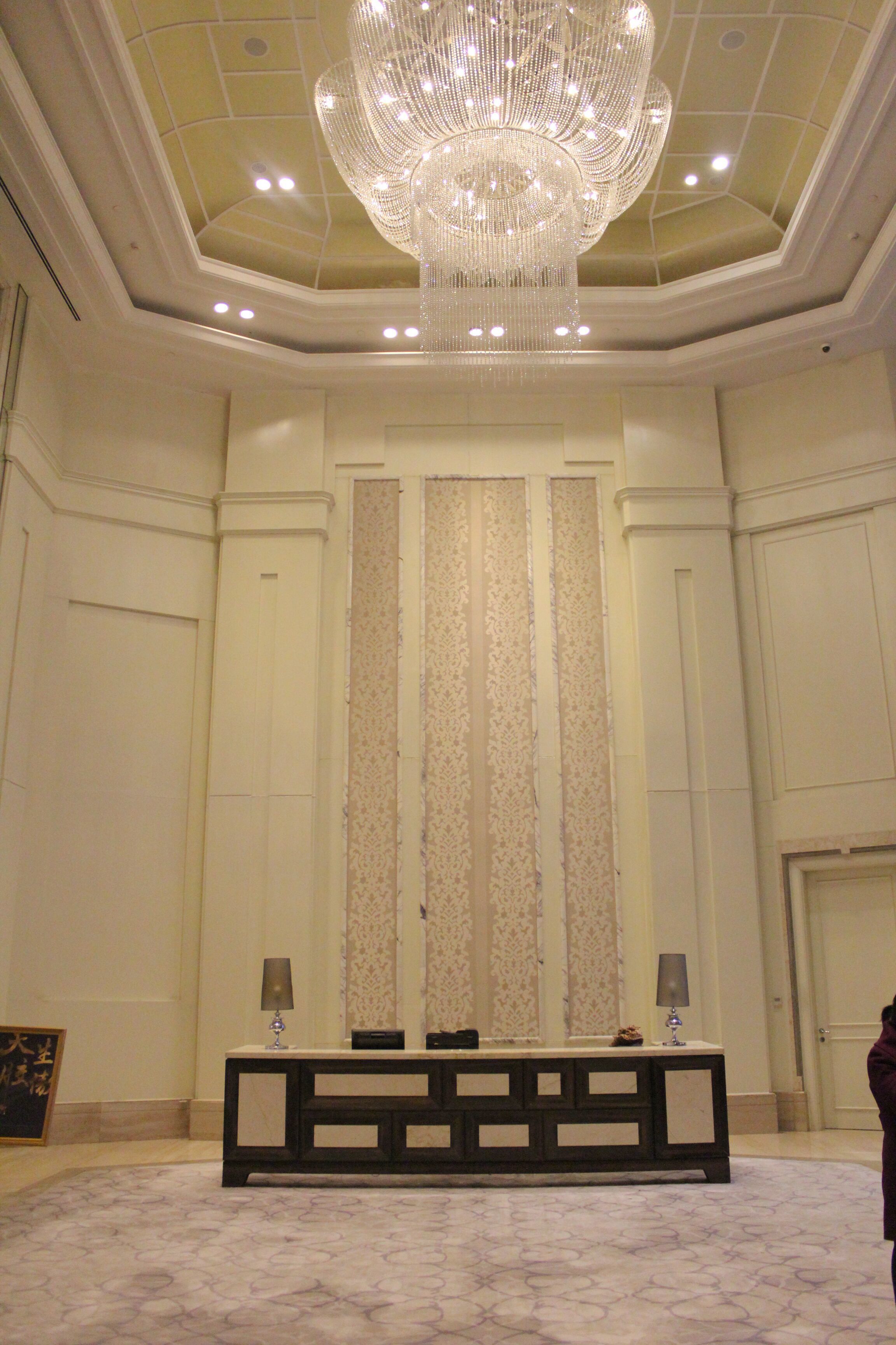Una sala elegante con un gran candelabro, una pared larga con paneles verticales y una consola con lámparas.