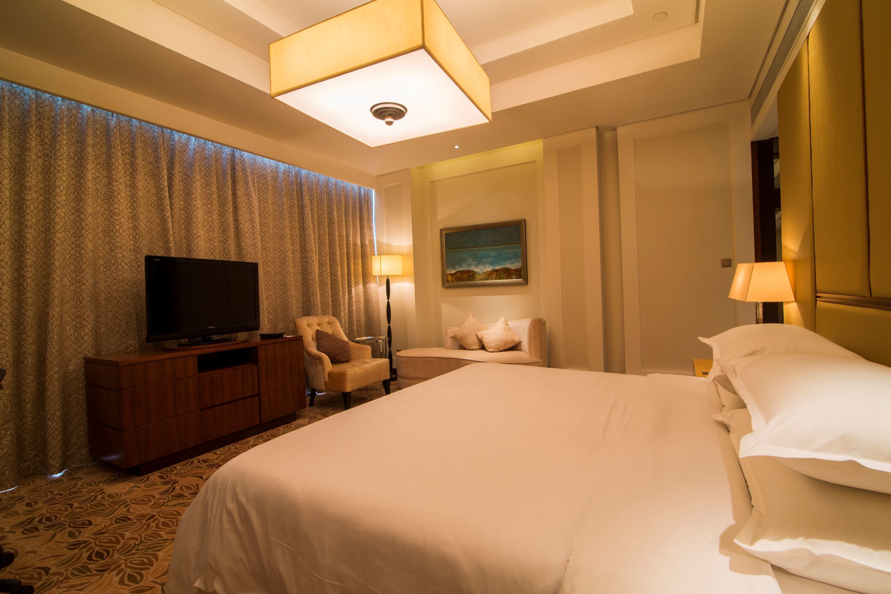 A hotel room with a large bed, a television on a wooden cabinet, a desk with a lamp, and a painting on the wall.