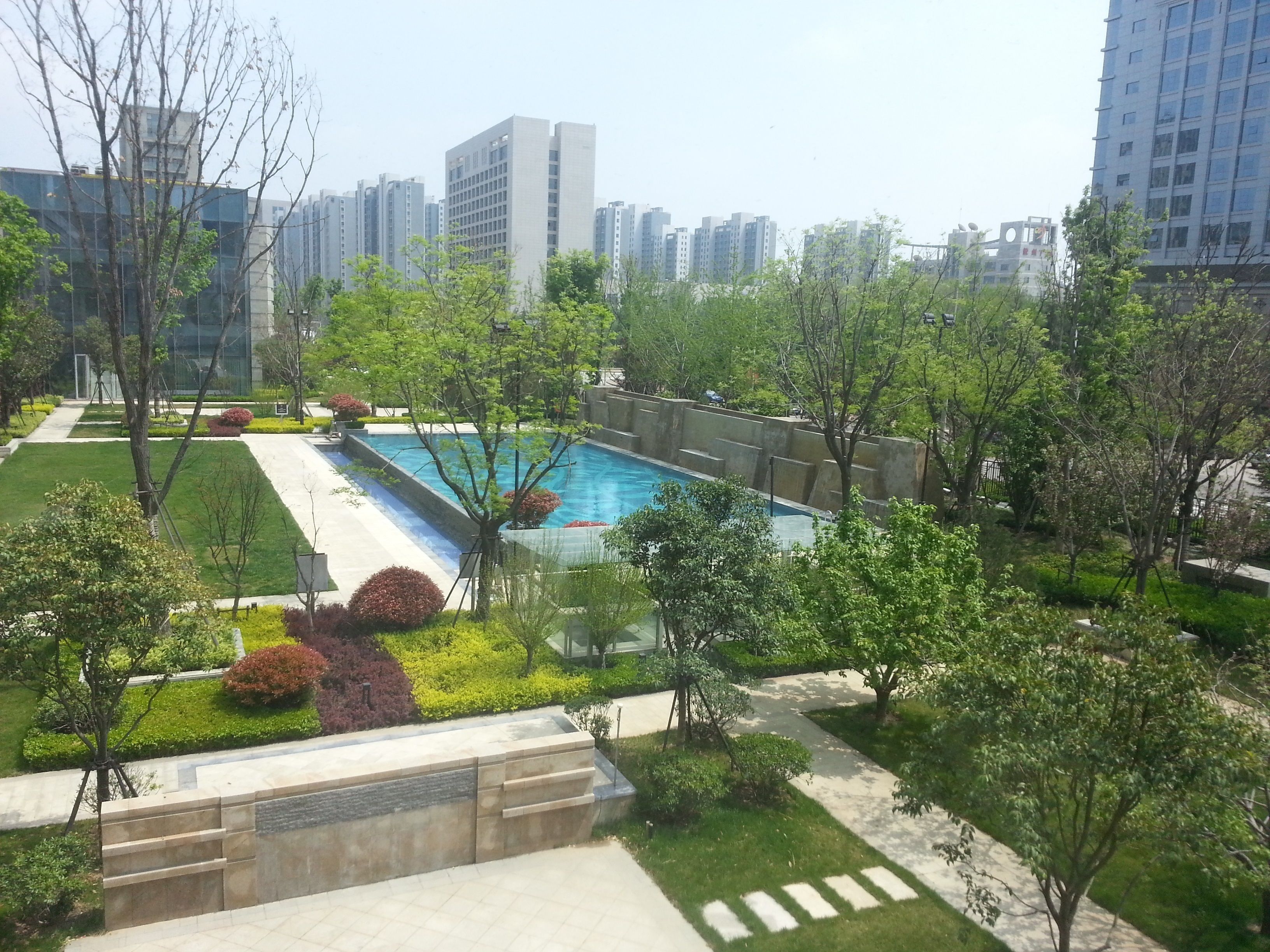A residential complex with a swimming pool surrounded by greenery and modern buildings in the background.