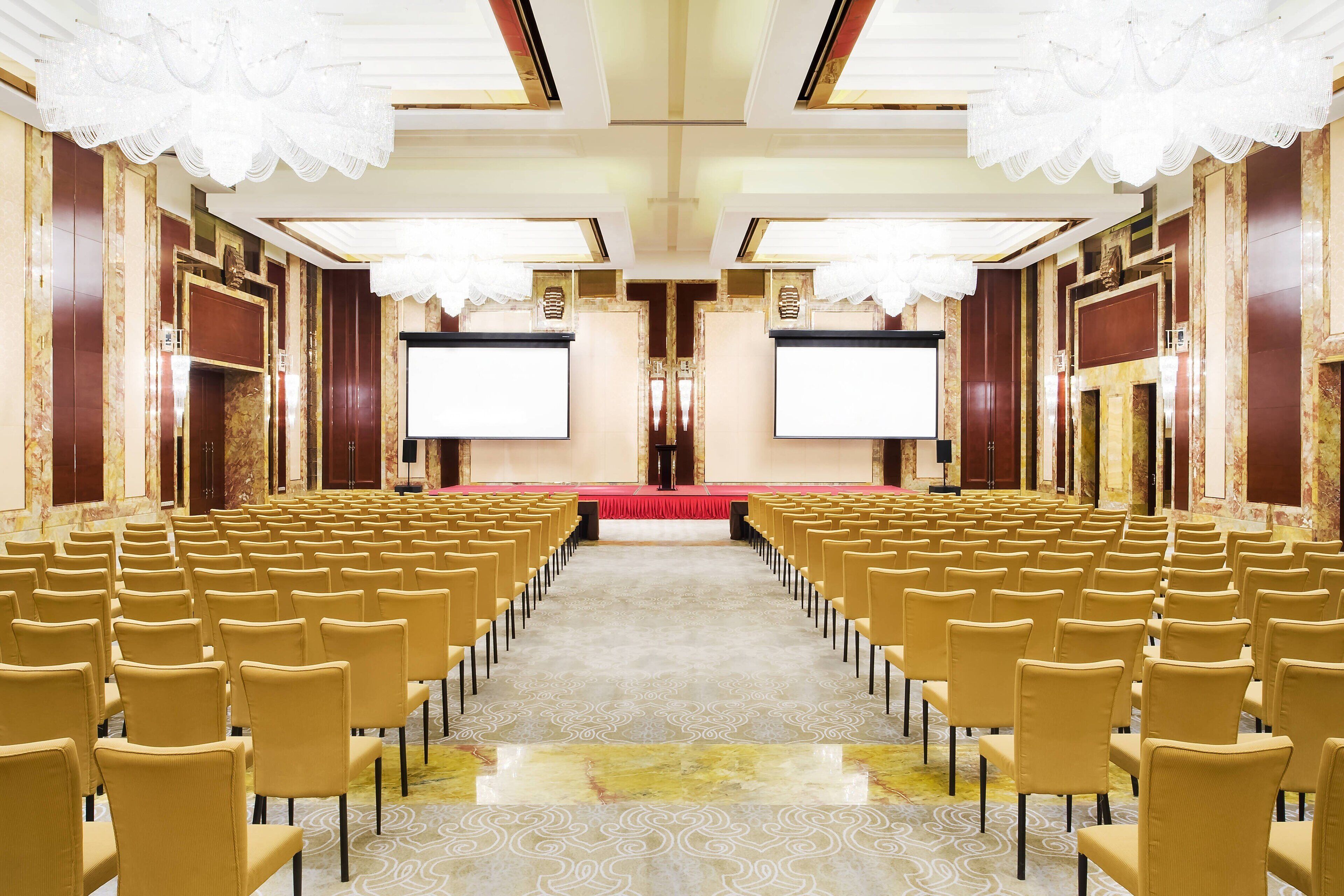 A conference room with rows of chairs facing a stage with a red carpet and two large screens.