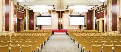 A conference room with rows of chairs facing a stage with a red carpet and two large screens.