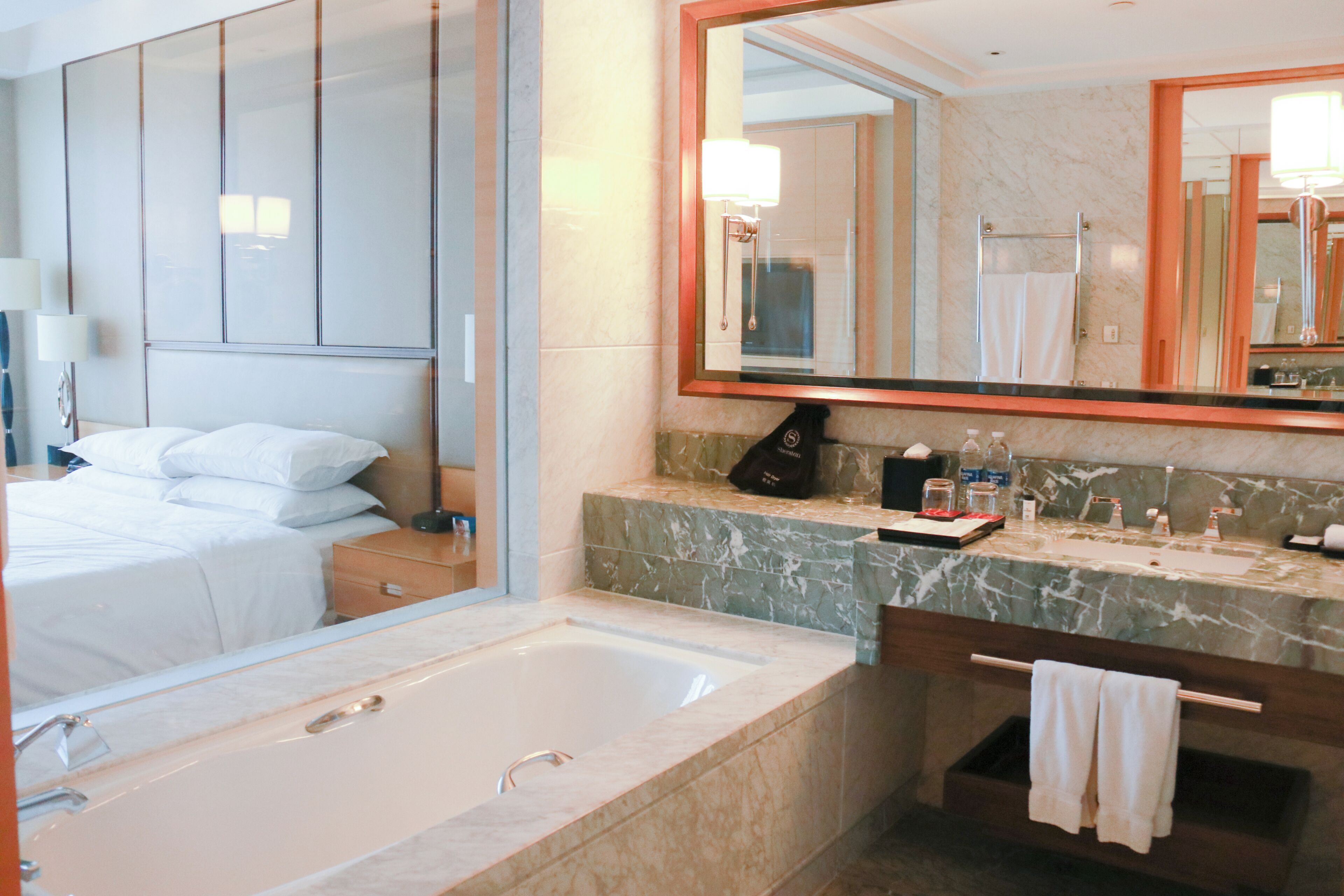A hotel bathroom with a large mirror, a bathtub, a vanity with a marble countertop, and a bed with white linens.