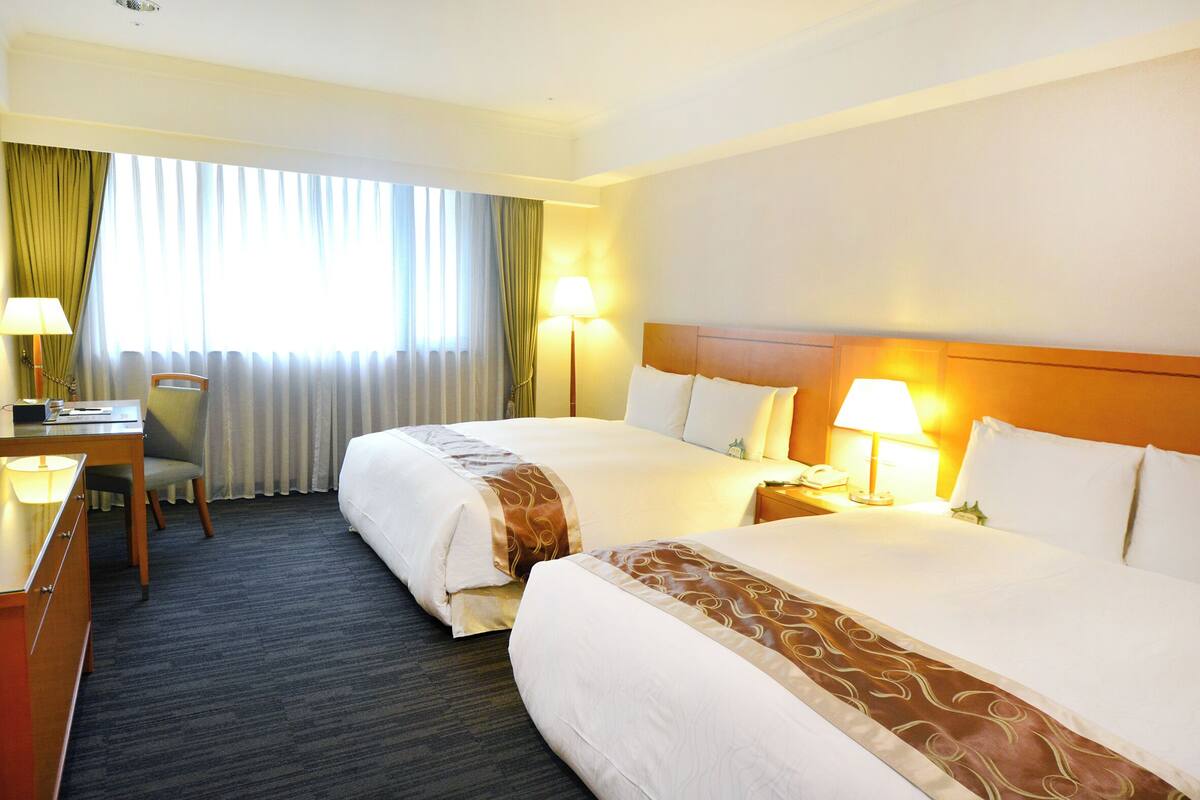 Une chambre d’hôtel avec deux lits, un bureau, une chaise et une fenêtre avec des rideaux.