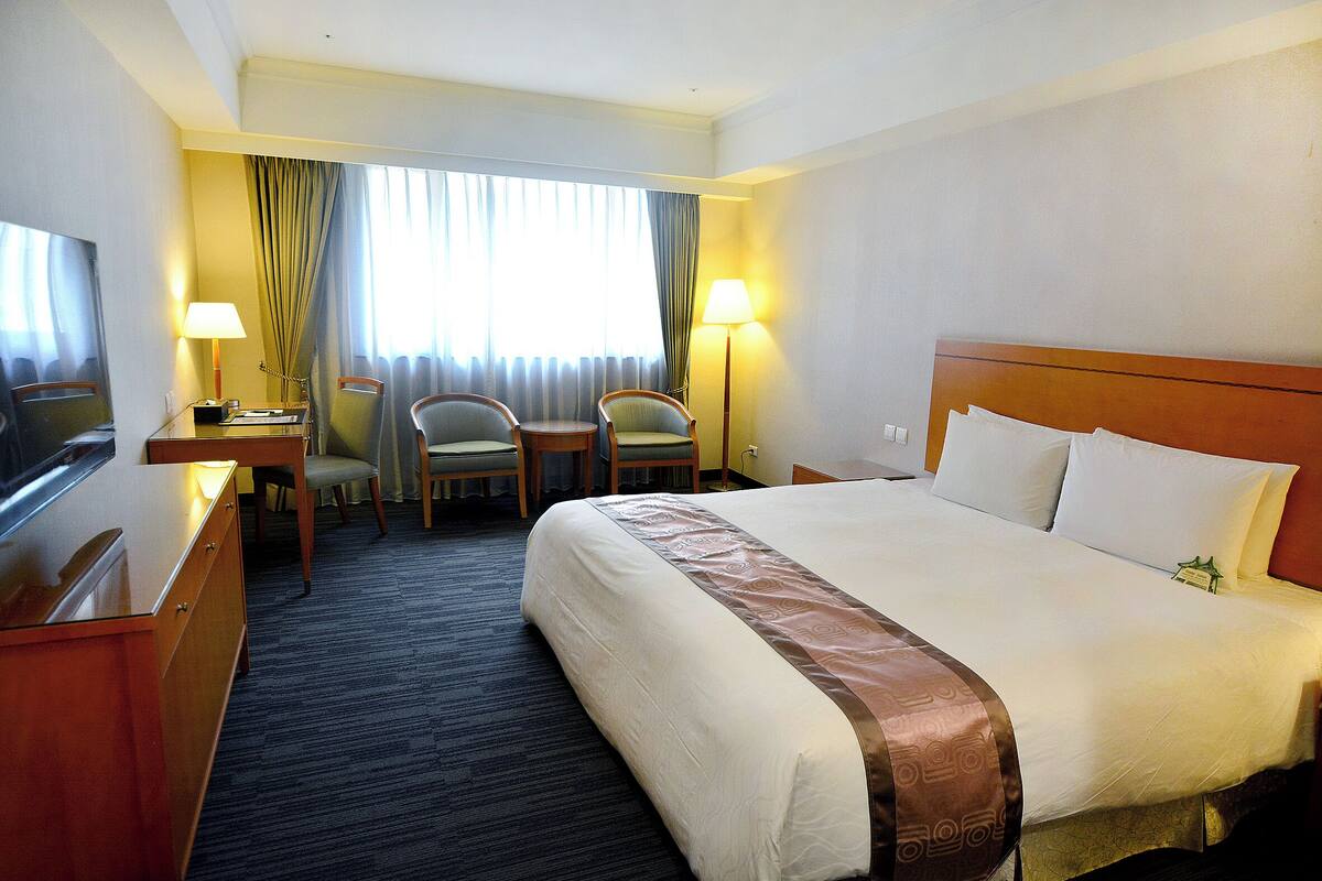 Une chambre d’hôtel avec un grand lit, un bureau avec une télévision, deux chaises, une petite table et une lampe.