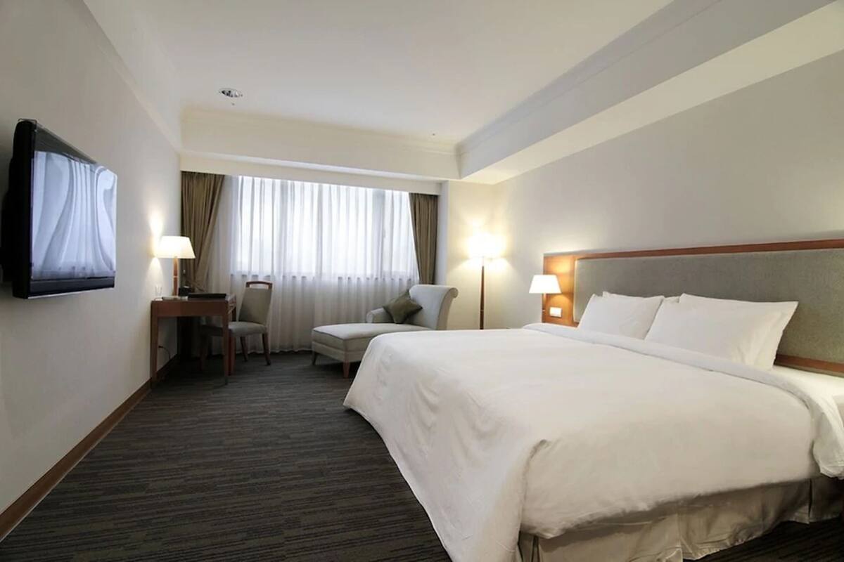 Une chambre d’hôtel avec un grand lit, un bureau avec une chaise, un téléviseur fixé au mur et une fenêtre avec des rideaux.
