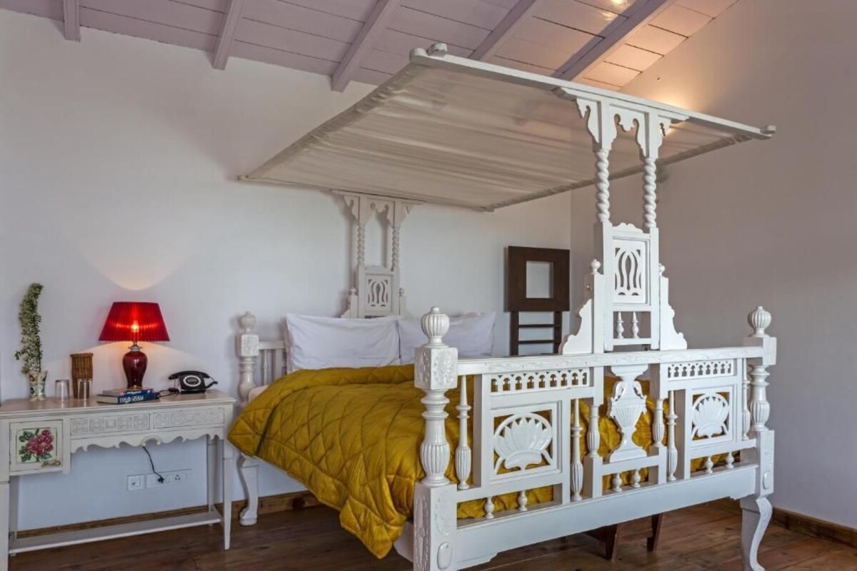 A white four-poster bed with a yellow bedspread, a bedside table with a lamp, and a mirror on the wall.