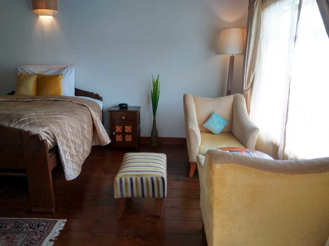 A bedroom with a bed, a chair, an ottoman, a nightstand, a lamp, and a plant.
