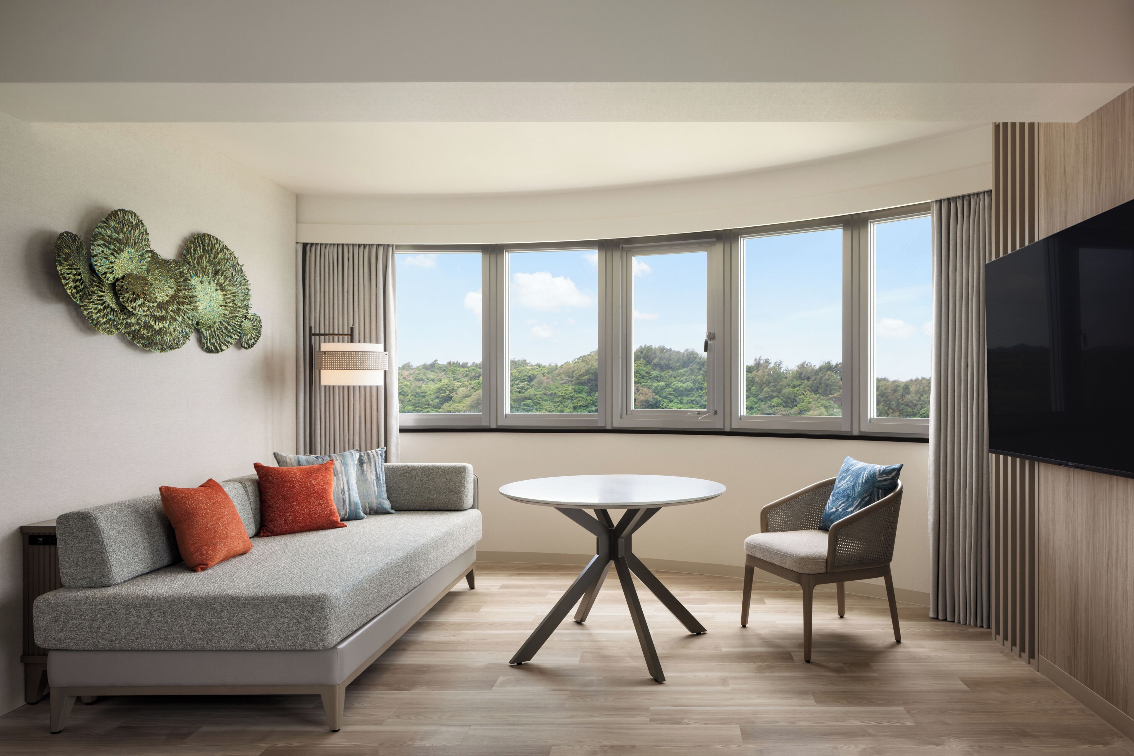 A modern living room with a sofa, armchair, round table, and large windows offering a view of greenery.