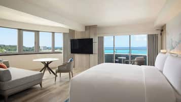 Habitación de hotel con cama, sofá, televisión, balcón con mesa y sillas, y ventanas amplias con vista al océano.
