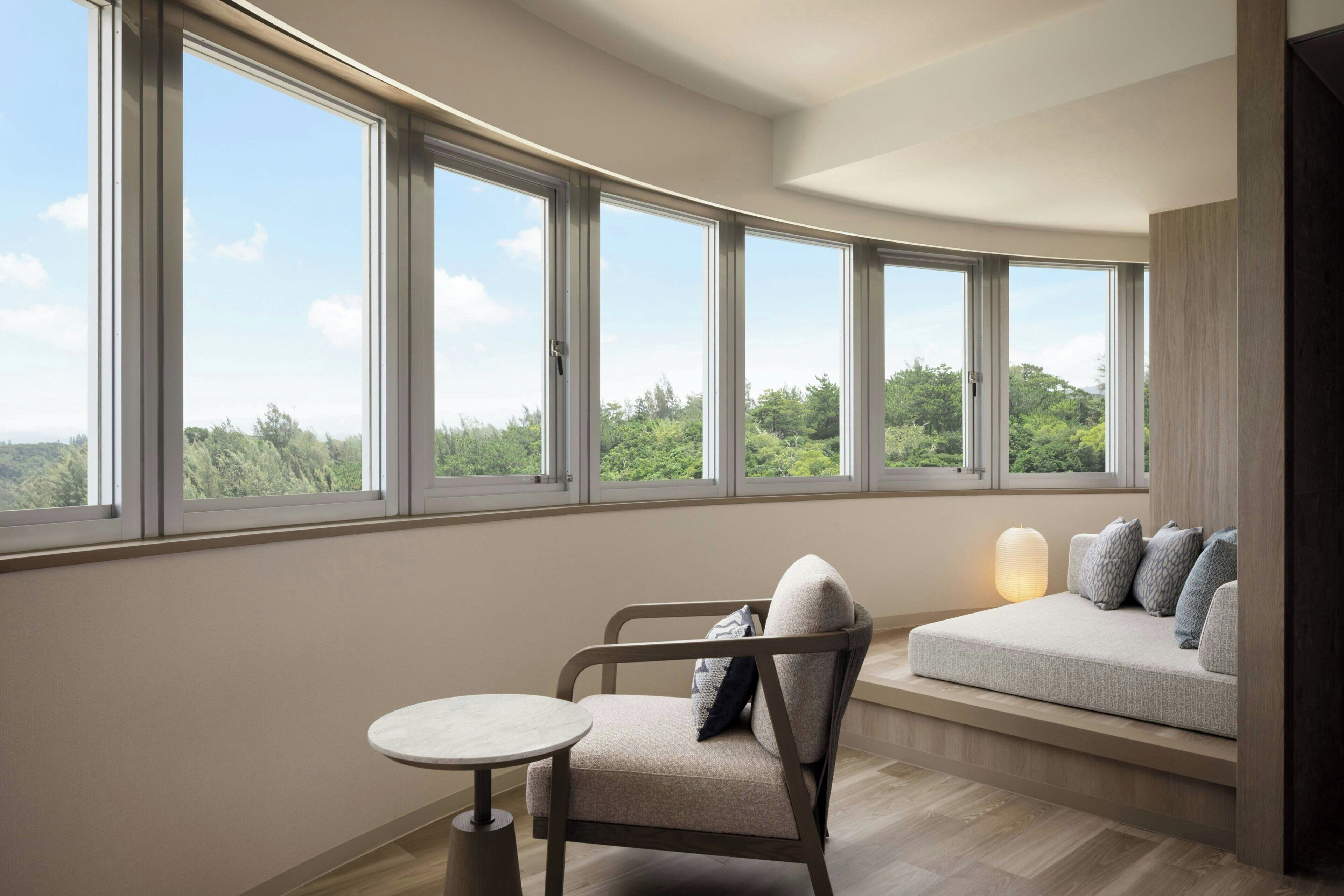 A room with a curved wall of large windows offering a view of greenery. There is a seating area with a cushioned bench and a small round table.