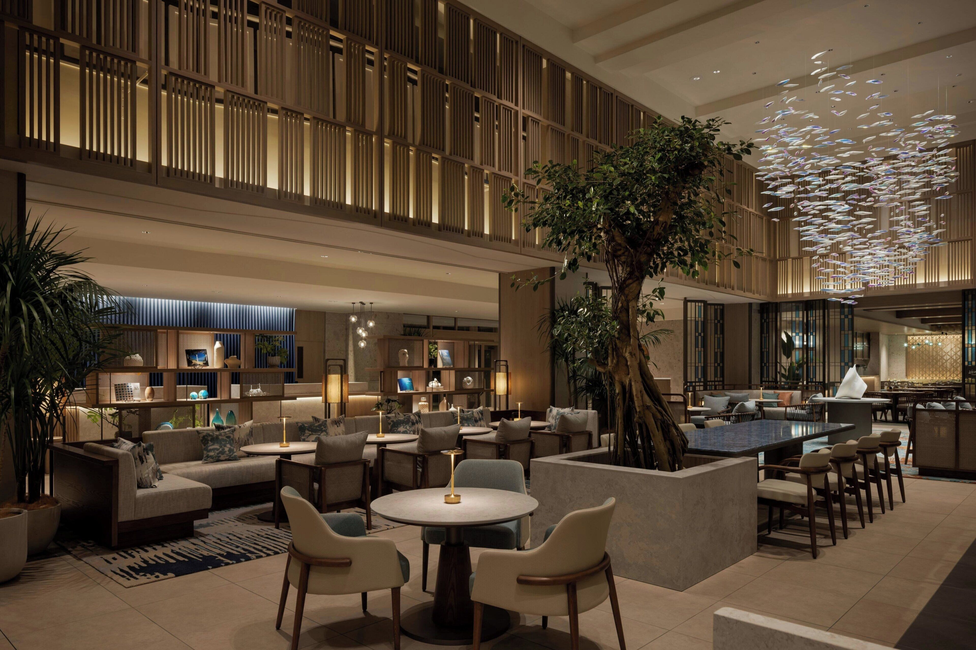A modern hotel lobby with a central seating area, a bar, and a large tree.