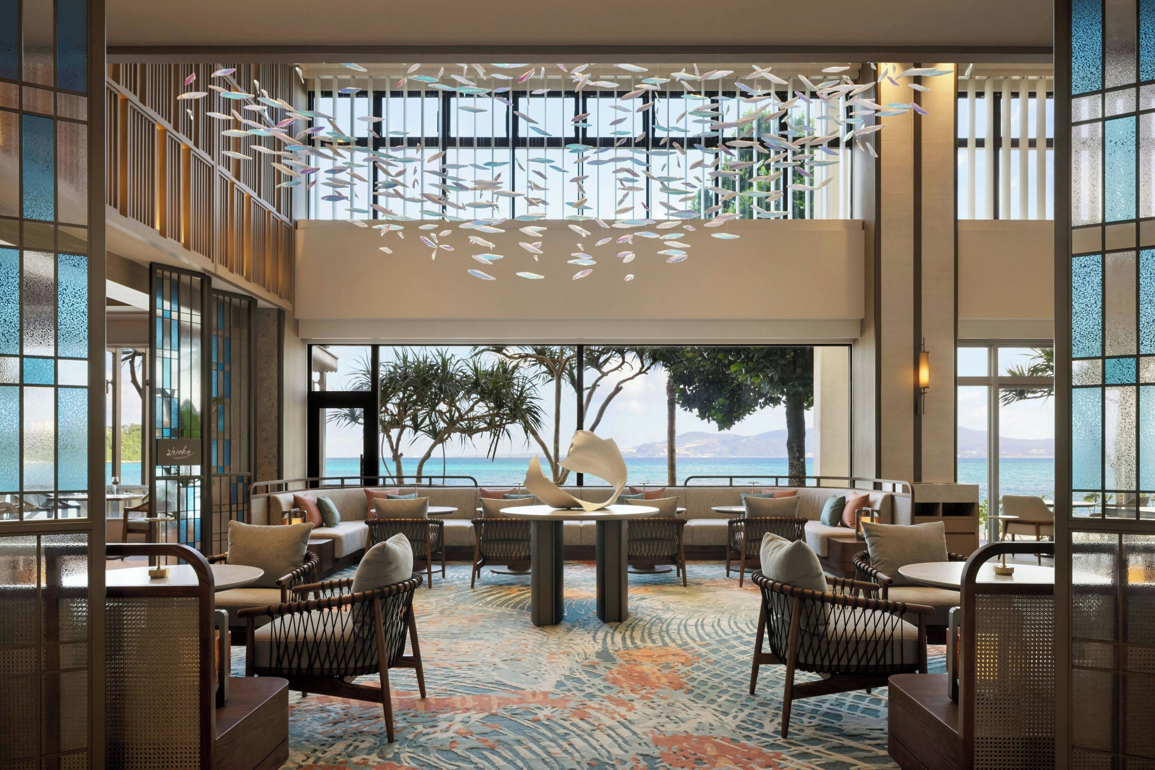 A spacious lounge with a high ceiling, a central table, and seating arranged around it. Large windows provide a view of the sea.