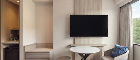 A hotel room with a TV, a round table, a sofa with a pillow, a small stool, and a minibar.