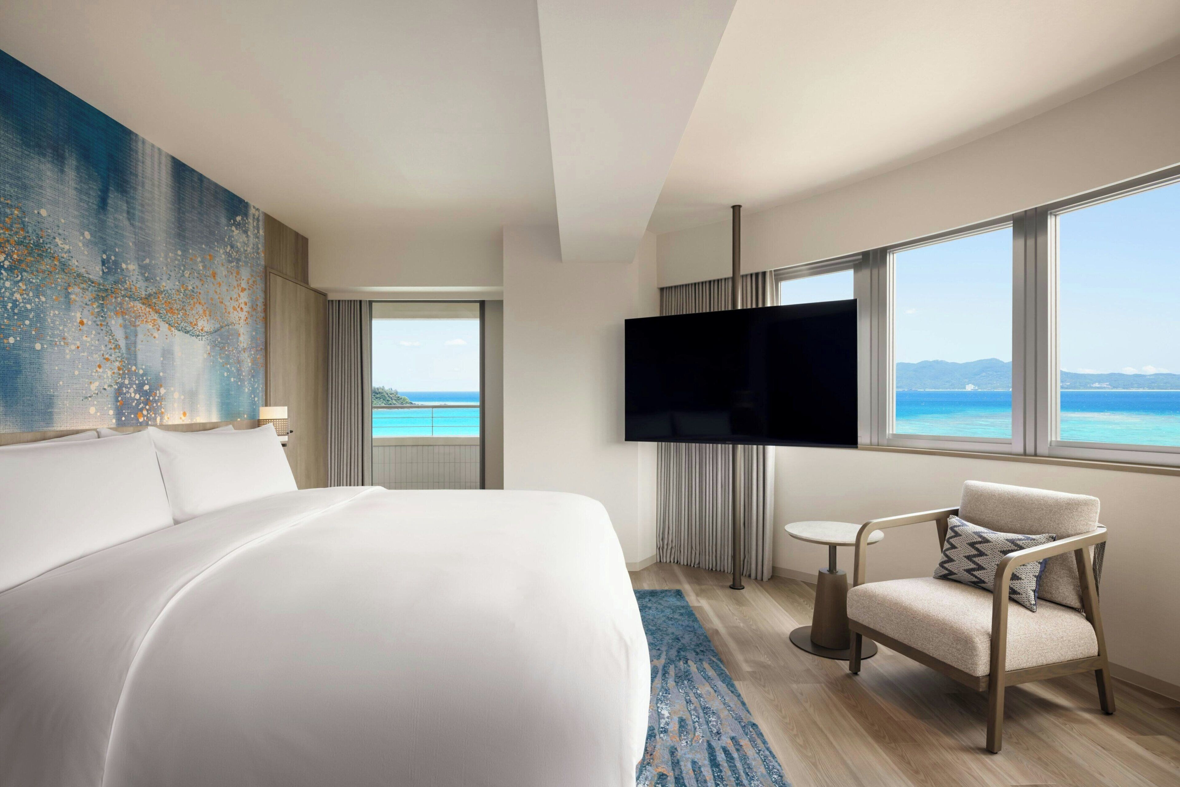 A hotel room with a bed, a TV, a chair, and a view of the ocean.