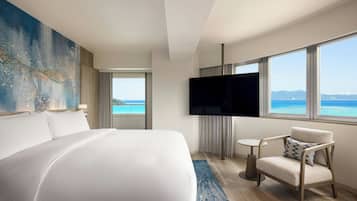 A hotel room with a bed, a TV, a chair, and a view of the ocean.
