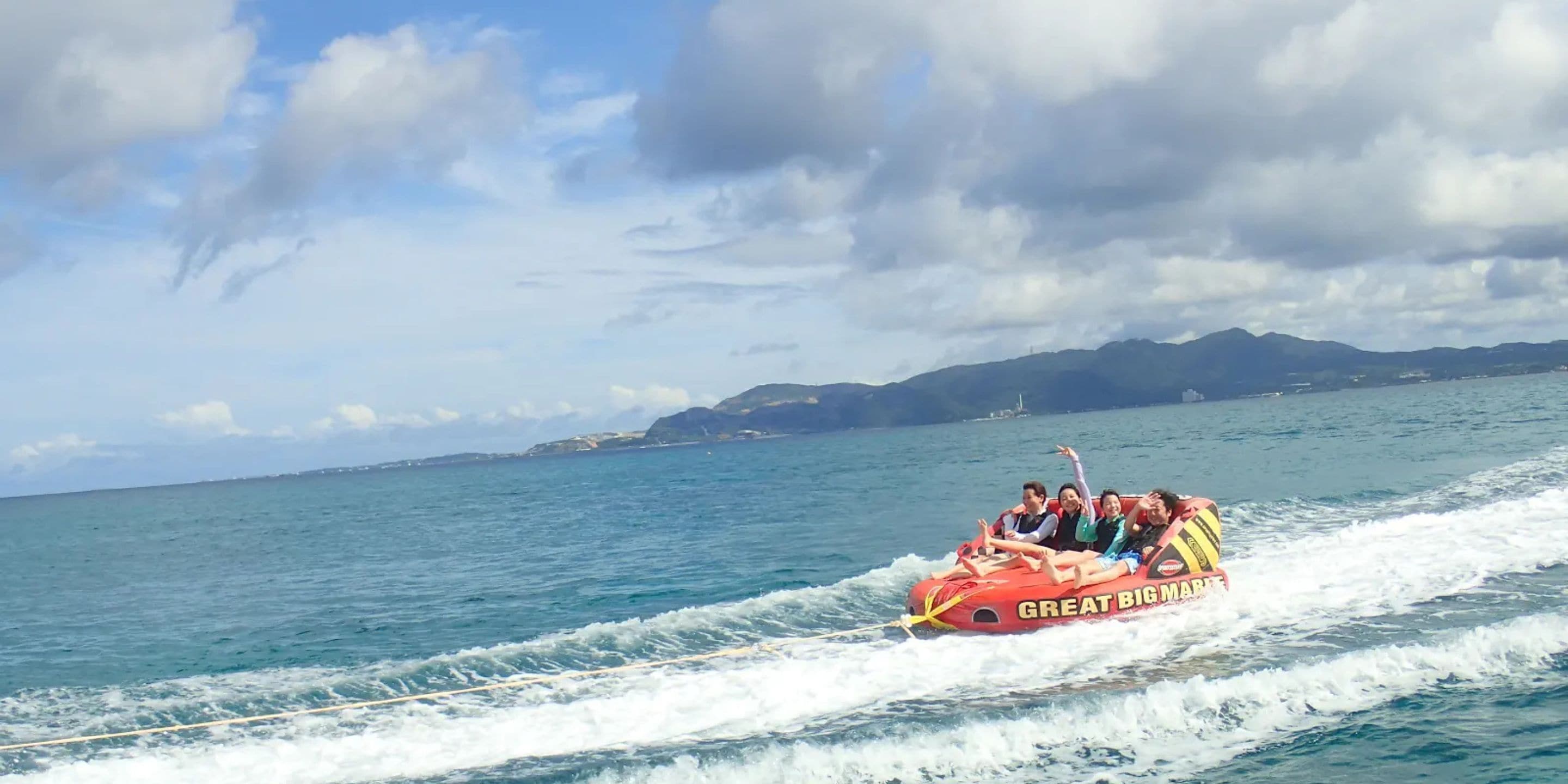 Een groep mensen op een rood vaartuig met de tekst 'GREAT AT BIG MARINE' erop, vaart over een wateroppervlak met een bergachtige kustlijn op de achtergrond.