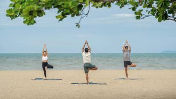 Três pessoas praticando yoga em uma praia arenosa, com o oceano ao fundo.