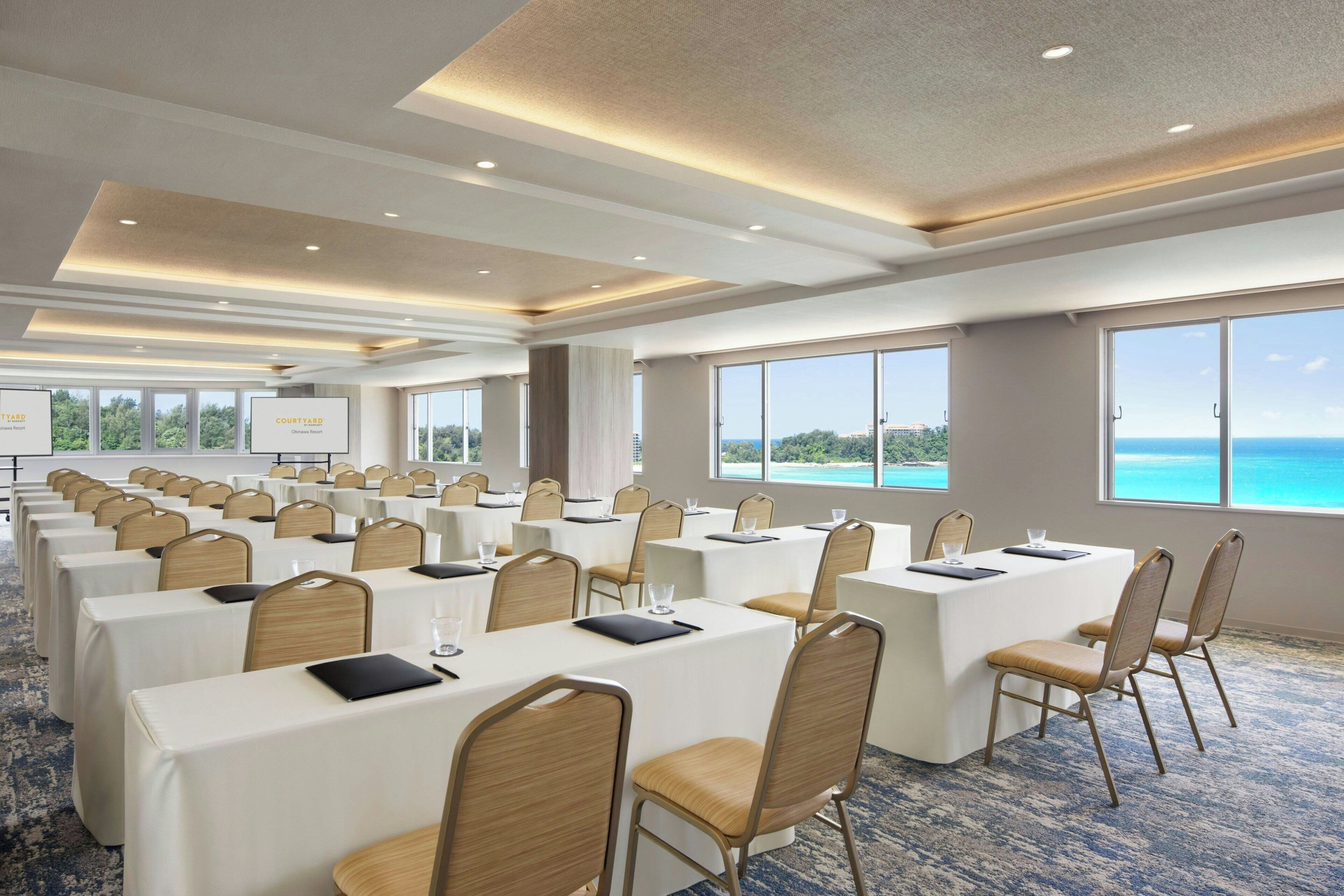 A conference room with multiple tables and chairs, large windows with a view of the ocean, and a presentation screen.
