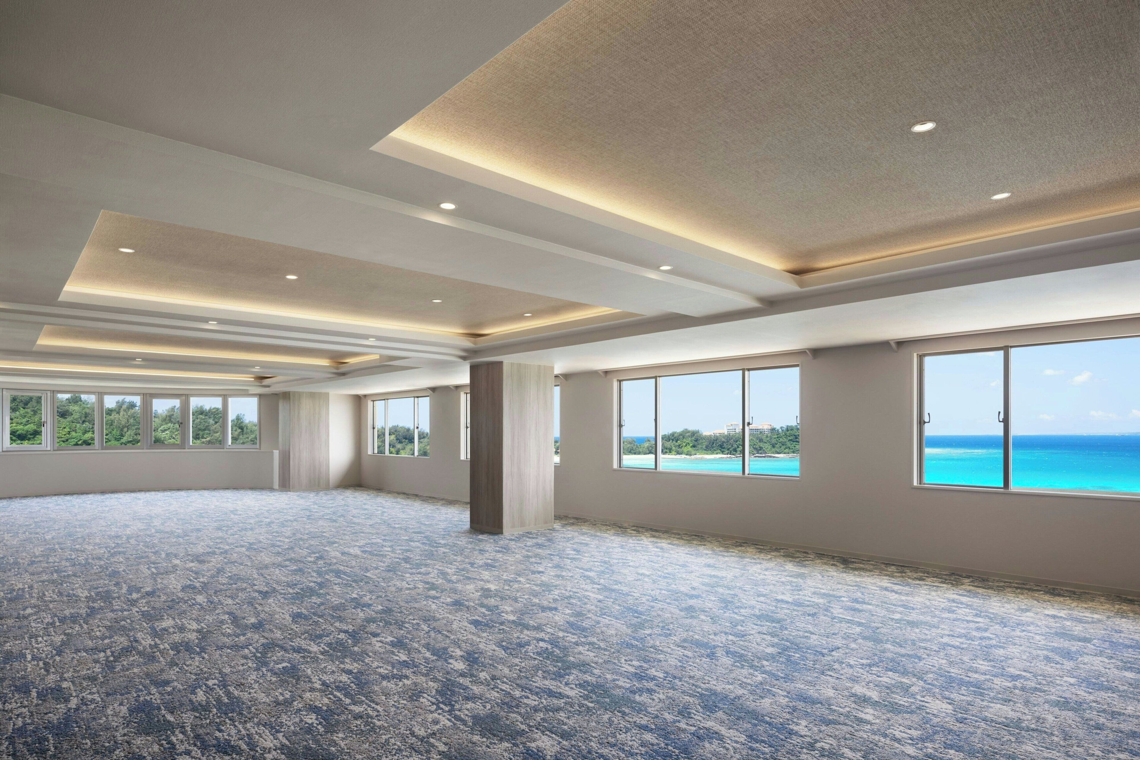 A large, open room with a patterned carpet, multiple windows with ocean views, and recessed ceiling lighting.